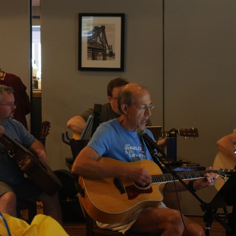 A guitarist singing into a microphone at an indoor jam, surrounded by other players.