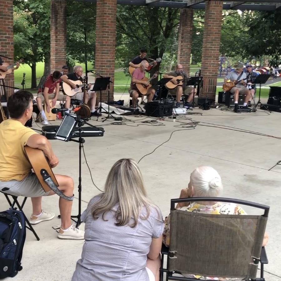 Musicians playing guitars together in a circle at an outdoor pavilion jam.